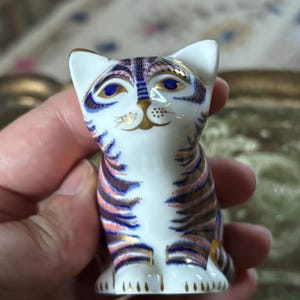 以下が含まれることがあります： 濃い青、ピンク、ゴールドの縞模様が手描きされた、白い磁器製の小さな猫の置物。鮮やかな青い目、金色の鼻、金色のひげが特徴で、光沢のある仕上げと繊細な模様が施されています。
