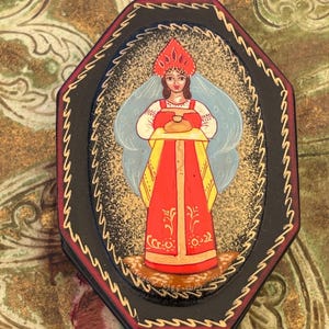 Peut inclure: Une boîte laquée octogonale noire ornée d'une illustration peinte à la main représentant une silhouette en sarafan rouge et coiffe kokoshnik tenant un pain. La bordure présente des motifs dorés et le texte "ЛЕВ-ШАЛ" à la base.