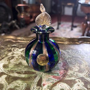 May include: A round glass perfume bottle featuring alternating vertical stripes of deep blue and emerald green, accented by thin gold-toned lines. The bottle has a clear, leaf-shaped glass stopper and a circular gold sticker labelled Bucella Cristalli Murano.