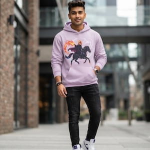 May include: A light purple hooded sweatshirt featuring a graphic print of a headless horseman riding a black horse with a flaming pumpkin head. The hoodie includes a front kangaroo pocket, long sleeves, and ribbed cuffs, paired with black jeans.