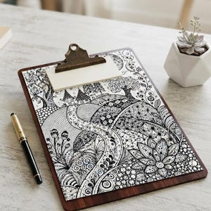 May include: A wooden clipboard holding a white sheet of paper with a detailed black and white ink illustration of a winding path, stylized trees, flowers, and small houses. A black and gold pen rests beside the clipboard on a light surface.