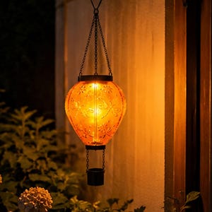 May include: A hanging lantern shaped like a hot air balloon with an orange glass body featuring embossed dragonfly and floral patterns. The lantern is suspended by black metal chains and includes a small black basket attached at the bottom.