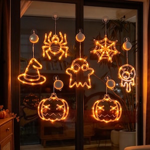 Może przedstawiać: Zestaw siedmiu pomarańczowych lampek LED na okno z motywami halloweenowymi: pająkiem, pajęczyną, kapeluszem czarownicy, duszkiem, gałką oczną i dwiema dyniami. Każda lampka jest przymocowana do przezroczystej przyssawki i małego białego pojemnika na baterie.