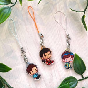 Peut inclure: Trois breloques en acrylique avec des illustrations de personnages de style chibi. Une breloque a un cordon bleu clair, une a un cordon orange et une a un cordon blanc. Chaque breloque comprend un fermoir mousqueton en métal et un anneau.
