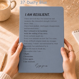 May include: A dusty blue notebook featuring the printed text I AM RESILIENT followed by a poem about overcoming hardship and the name Syazana at the bottom. The cover has a smooth matte finish and the text is printed in a clear black sans-serif font.