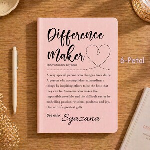 Puede incluir: Un cuaderno rosa claro con el texto negro "Difference Maker" y un contorno de corazón punteado. Debajo, una definición al estilo de diccionario describe a una persona que cambia vidas, seguida del texto "See also: Syazana" en negro.