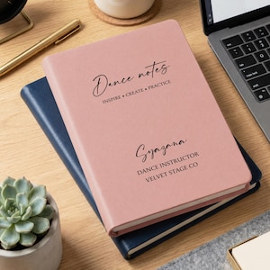 Könnte beinhalten: Ein rosa Notizbuch mit den Worten "Dance notes" und "INSPIRE • CREATE • PRACTICE" auf dem Einband. Darunter stehen der Name "Syazana" und der Text "DANCE INSTRUCTOR VELVET STAGE CO". Das Notizbuch liegt auf einem blauen Notizbuch.