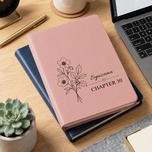 May include: A dusty pink hardcover journal featuring a black line art illustration of a floral sprig with holly leaves and berries. The text Syazana and CHAPTER 30 are printed in black ink on the front cover, separated by a small heart icon.