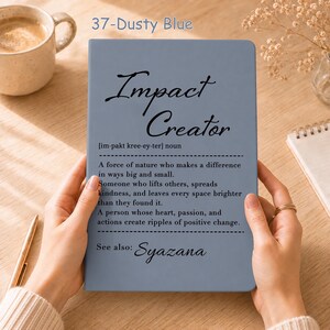 Puede incluir: Un cuaderno azul polvoriento con el texto impreso en negro Impact Creator, una pronunciación fonética y una definición que describe a una persona que marca la diferencia, con el nombre Syazana bajo el encabezado See also.