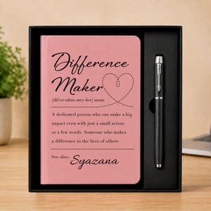 Op de afbeelding: Een roze notitieboek en een zwarte pen in een zwarte geschenkdoos. De kaft van het notitieboek bevat de tekst Difference Maker, een fonetische uitspraak, een definitie van de term en de naam Syazana, alles in het zwart met een gestippelde hartvorm.