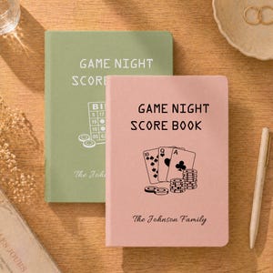 Könnte beinhalten: Ein salbeigrünes Notizbuch mit dem Titel GAME NIGHT SCORE und einer Bingo-Grafik sowie ein altrosa Notizbuch mit dem Titel GAME NIGHT SCORE BOOK mit einer Grafik aus Spielkarten und Pokerchips, beide mit dem Text The Johnson Family.