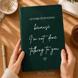 May include: A dark forest green journal with white text that reads LETTERS TO SYAZANA, because I'm not done talking to you, and a small heart icon. The cover has a smooth, matte finish and is held by two hands against a wooden surface.