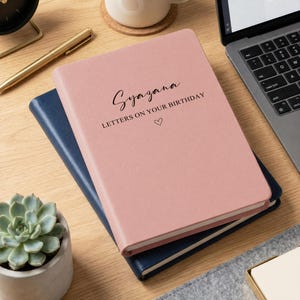 Op de afbeelding: Een oudroze notitieboek met harde kaft waarop de tekst Syazana LETTERS ON YOUR BIRTHDAY en een klein hartje in het zwart op de voorkant staan gedrukt. Het notitieboek ligt op een donkerblauw schrift naast een gouden pen en een laptop.