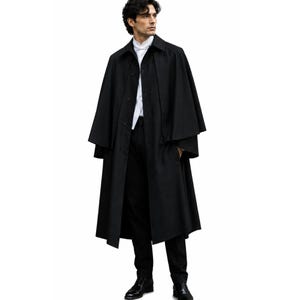 Op de afbeelding: Een lange zwarte trenchcoat met een cape-achtig ontwerp. De jas is dichtgeknoopt en wordt gedragen over een wit overhemd met kraag en een zwarte broek. Het model draagt zwarte schoenen.