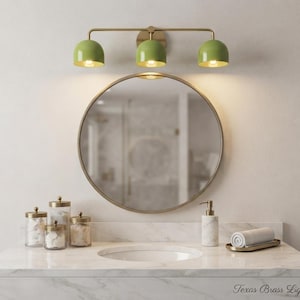 Può includere: Lampada da bagno a tre luci con finitura in ottone e paralumi a cupola verde salvia montata sopra uno specchio circolare. Il piano in marmo include barattoli in vetro, un dispenser di sapone in marmo e un asciugamano arrotolato. Testo: Texas Brass Lights.