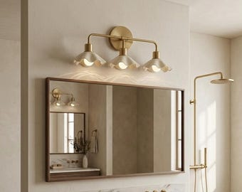 Puede incluir: Una lámpara de pared de tres luces con acabado en latón cepillado y pantallas de cerámica festoneadas en color blanco roto. La lámpara está montada sobre un espejo rectangular con marco de madera oscura que refleja un baño con ducha.
