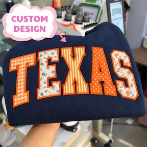 May include: A navy blue sweatshirt featuring the word TEXAS in large block letters. Each letter has a bright orange border and a unique pattern, including orange gingham, blue floral, orange stripes, orange polka dots, and red floral prints.