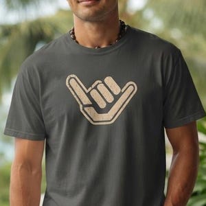 May include: A charcoal gray short-sleeve t-shirt featuring a beige shaka hand sign graphic centered on the chest. The shirt has a crew neckline and a standard fit, showcasing a simple design with a neutral color palette and a clear hand gesture print.