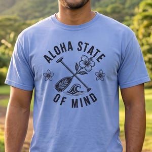 May include: A light blue short-sleeve t-shirt featuring a black graphic print of a paddle, a hibiscus flower, and ocean waves. The text "ALOHA STATE OF MIND" is printed in a circular arrangement around the central graphic design.
