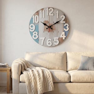 May include: A large, round wall clock with a distressed, rustic design. The clock face features white numerals against a light wood background with blue and brown accents. Black clock hands indicate the time. The clock is mounted on a white wall.