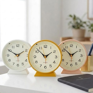Puede incluir: Tres relojes de cuarzo analógicos en blanco, amarillo y rosa. Cada reloj tiene una esfera redonda con manecillas negras, un segundero y una esfera blanca con números negros. Los relojes están sobre una superficie blanca.