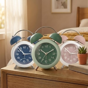 May include: Three round twin-bell alarm clocks in blue, green, and pink. Each clock features a white frame, a matching colored face with white Arabic numerals, silver hands, and a quartz movement. The clocks stand on small silver metal legs.