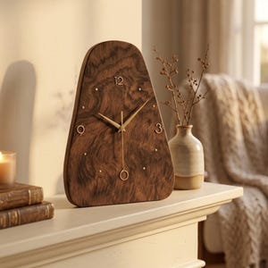 May include: A teardrop-shaped wooden table clock with a dark brown wood grain finish. The clock face features gold-colored hands and gold-colored numerals at the 12, 3, 6, and 9 o'clock positions, with small gold dots marking the remaining hours.