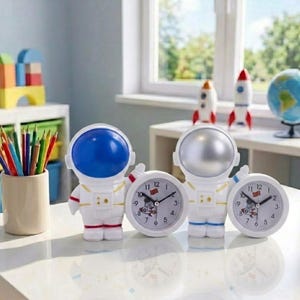May include: Two astronaut-themed analog clocks with white suits and gold accents. One astronaut has a blue helmet, the other a silver helmet. Each clock face features black numbers, hour and minute hands, and a small graphic of a rocket ship.