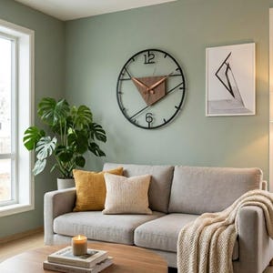 May include: A large circular wall clock with a black metal frame, featuring a central triangular wooden accent and copper-colored hands. The clock displays the numbers 12 and 6 in a black font, mounted on a sage green wall above a light gray sofa.
