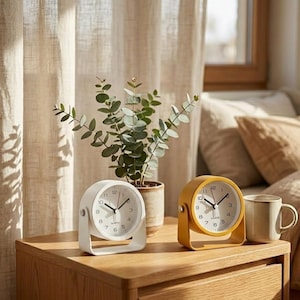May include: Two analog alarm clocks, one white and one yellow, sit on a wooden nightstand. The white clock has a curved frame, while the yellow clock has a rectangular frame. A small ceramic mug and a potted plant are also on the nightstand.