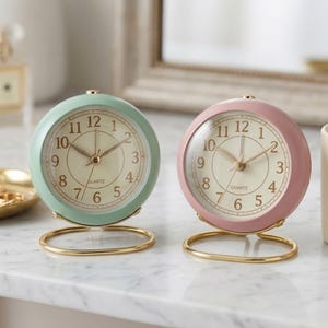 May include: Two small, round alarm clocks. One is mint green, the other is pink. Both have a gold-colored base and hands, with a cream-colored clock face. The clock face has black numbers and the word "QUARTZ".
