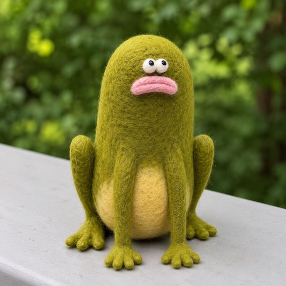A hand-needle-felted sad cucumber frog is pure chaotic desk energy — someone sculpted this tiny melancholy amphibian by hand and it shows.
