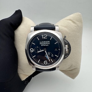 Pode incluir: Um relógio Panerai Luminor BiTempo em aço inoxidável com mostrador preto e bracelete em couro preto. O relógio tem um mostrador quadrado com algarismos e ponteiros brancos e um pequeno mostrador de segundos. O relógio é exibido em uma almofada creme.