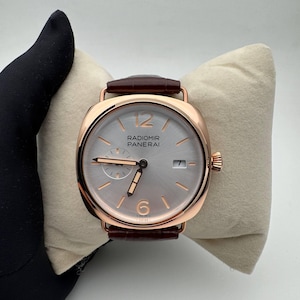 Pode incluir: Um relógio Radiomir Panerai em ouro rosa com uma pulseira de couro castanho. O relógio tem um mostrador prateado com ponteiros e algarismos em ouro rosa. O relógio é exibido em uma almofada bege.