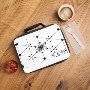 Peut inclure: Un sac à déjeuner rectangulaire avec un extérieur blanc orné d'un motif géométrique de flocon de neige noir et du texte "BY S DESIGNS" dans un cadre rectangulaire. Le sac possède une fermeture éclair noire et une poignée supérieure noire.