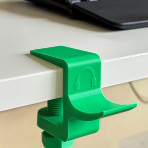 May include: A bright green 3D-printed headphone hanger clamp featuring a curved cradle for resting headphones. The device includes a threaded screw mechanism for securing it to a desk edge and displays a small embossed icon of headphones.