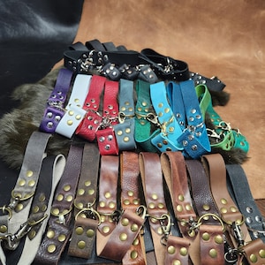 May include: A collection of leather keychains with metal rings and decorative studs. The keychains are in various colors, including black, white, red, blue, green, brown, and tan.