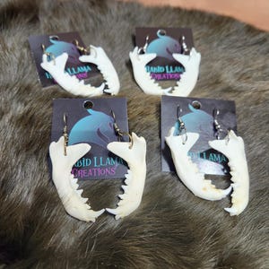 May include: Four pairs of white bone jaw earrings on cards with the text "Rabid Llama Creations".