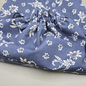Könnte beinhalten: Ein blauer Beutel mit Kordelzug und einem weißen sowie gelben Blumenmuster. Die Stofftasche hat einen gerafften Verschluss, der mit dünnen schwarzen Kordeln gesichert ist, und zeigt einen durchgehenden botanischen Druck auf der gesamten Oberfläche.