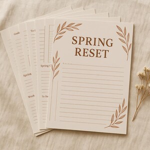 Pode incluir: Uma pilha de blocos de notas creme com a frase "SPRING RESET" em castanho. Os blocos têm páginas pautadas e detalhes de folhas. Um raminho de flores secas está ao lado.