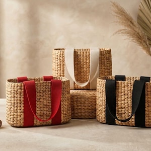 May include: Three rectangular woven straw baskets with flat bottoms and rounded corners. Each basket features two sturdy fabric handles in solid colors: one set is bright red, one set is cream, and one set is black. The weave is uniform and natural.