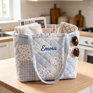 May include: A quilted tote bag featuring a light blue and white patchwork pattern with floral and gingham squares. The name Emma is embroidered in blue script on the front. The bag contains a white towel, a small case, and sunglasses.
