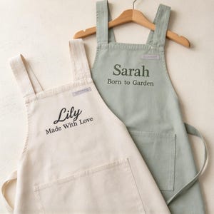 Op de afbeelding: Twee schorten hangen aan een houten kledinghanger. De ene is crème met de geborduurde tekst Lily Made With Love. De andere is saliegroen met de geborduurde tekst Sarah Born to Garden. Beide schorten hebben brede schouderbanden en een voorzak.