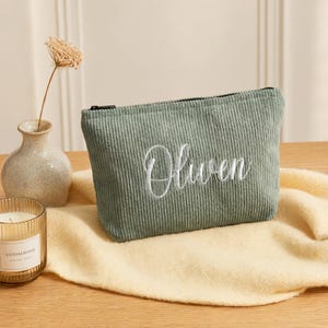May include: A sage green corduroy zippered pouch featuring the name Oliven embroidered in white cursive script. The pouch has a textured ribbed fabric surface and a black zipper closure along the top edge, presenting a simple rectangular shape.