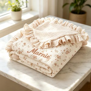 May include: A cream-colored muslin blanket featuring a delicate brown floral pattern and a ruffled edge. The name Marnie is embroidered in brown script on the front of the folded fabric, which has a textured, multi-layered cotton appearance.