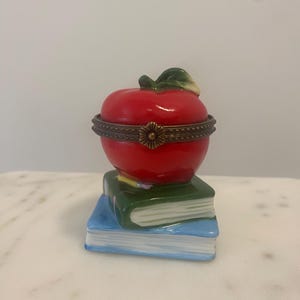 May include: A porcelain trinket box shaped like a bright red apple with a green leaf, resting on a stack of two books. The top book is dark green and the bottom book is light blue. A decorative brass metal hinge with a flower clasp connects the apple.