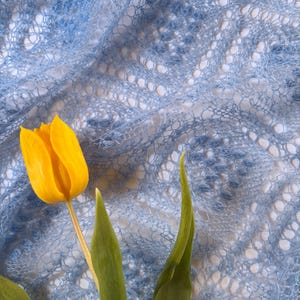 Könnte beinhalten: Eine leuchtend gelbe Tulpe mit grünen Blättern vor einem hellblauen, offen gewebten Textil. Das Textil hat ein zartes, spitzenartiges Muster. Das Bild ist eine Nahaufnahme, die die Texturen und Farben hervorhebt.