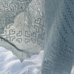May include: A light blue, intricately knitted shawl with a delicate lace pattern. The shawl features a scalloped edge and heart-shaped designs, creating a soft, airy texture. The shawl is displayed against a snowy background.