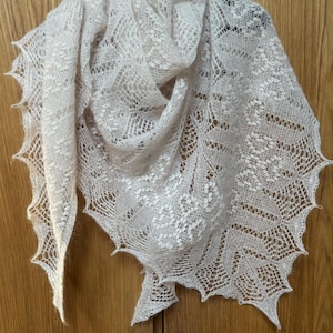May include: A light beige, open-weave shawl with a delicate, lacy pattern. The shawl is draped over a wooden surface, showcasing its intricate design and scalloped edges. The soft color and texture suggest a lightweight, comfortable accessory.