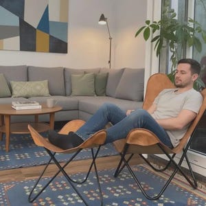 May include: A tan leather butterfly chair with a matching ottoman, both featuring a curved sling seat design supported by a black metal frame. The chair and ottoman are positioned on a blue patterned rug in a living room setting.